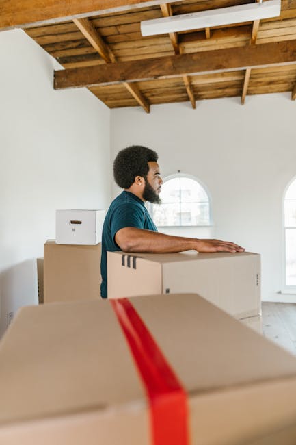 A person with visible arm tattoos and wearing a blue t-shirt and grey trousers is inside a room, holding a cardboard box over their head with one hand for support and walking near a window. The room h