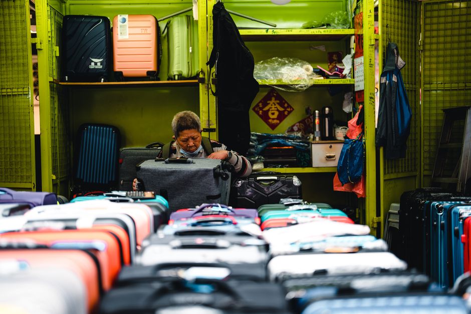 A woman organizing and packing suitcases and bags inside a small storage area or shop, with metal shelving units filled with suitcases, backpacks, and plastic-wrapped items behind her. The suitcases i
