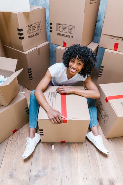 A woman with curly black hair, smiling and sitting on a wooden floor surrounded by large cardboard moving boxes, some sealed with red tape and others open with packing materials visible. She is wearing a white t-shirt, jeans, and white sneakers, holding a sealed box with her right hand. The background shows a wall with peeling blue paint, indicating an indoor setting during a home relocation process. The scene captures the packing and moving stage, with boxes ready for transport, reflecting the logistics involved in furniture transport and home removals. Movers Brixton is providing professional house removal services, focusing on efficient and careful loading procedures in tight access areas, as suggested by the presence of multiple packed items and moving equipment such as boxes and packaging materials.