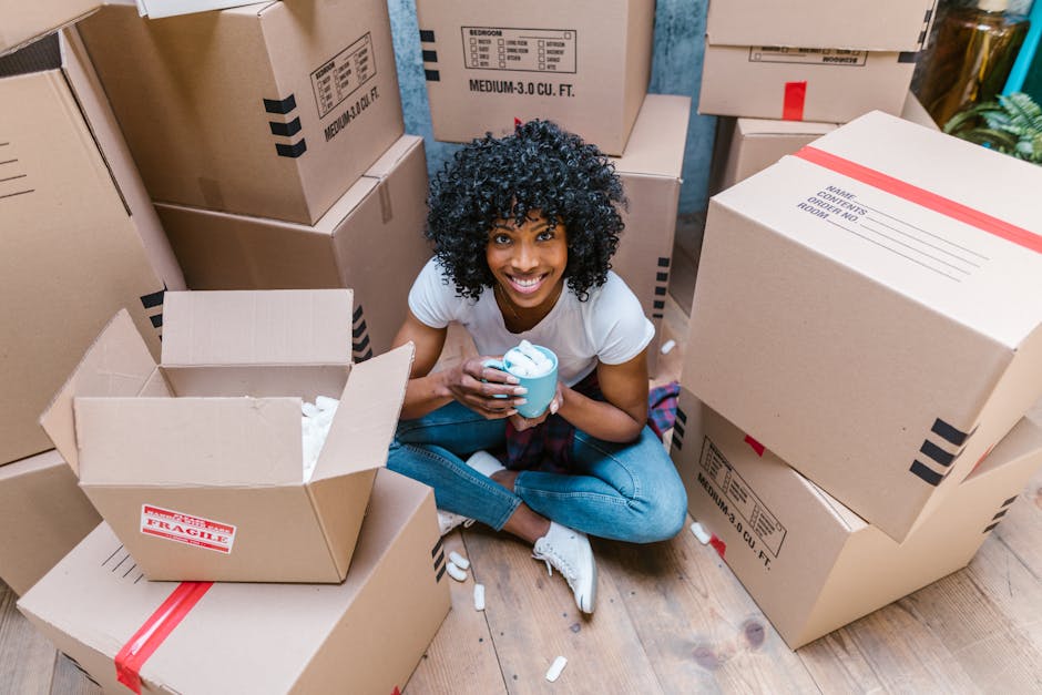 A young woman with curly black hair, wearing a white t-shirt and blue jeans, is sitting on a wooden floor surrounded by stacked cardboard boxes of various sizes during a home relocation. Some boxes are sealed with red or black tape, while others are open, revealing packing materials like tissue paper and protective padding. She is holding a blue cup and smiling towards the camera, and crumpled tissue paper and packing fillers are scattered around her. Behind her, additional boxes are stacked against a light blue wall, and a few indoor plants can be seen to the right. The scene depicts the moving process, including packing and preparing items for transportation, illustrating the logistical aspect of a house move as managed by [COMPANY_NAME], which offers professional removals and furniture transport services, especially when dealing with tight access situations during home relocation near Brixton.