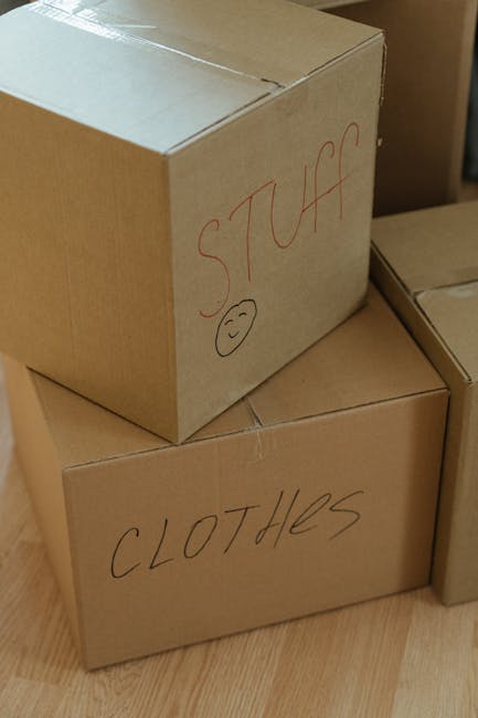 A woman with curly black hair, smiling and sitting on a wooden floor surrounded by large cardboard moving boxes, some sealed with red tape and others open with packing materials visible. She is wearing a white t-shirt, jeans, and white sneakers, holding a sealed box with her right hand. The background shows a wall with peeling blue paint, indicating an indoor setting during a home relocation process. The scene captures the packing and moving stage, with boxes ready for transport, reflecting the logistics involved in furniture transport and home removals. Movers Brixton is providing professional house removal services, focusing on efficient and careful loading procedures in tight access areas, as suggested by the presence of multiple packed items and moving equipment such as boxes and packaging materials.
